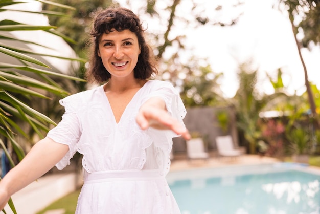 Feliz jovem morena caucasiana em vestido de verão sorri e alcança a câmera com as mãos ao ar livre no verão