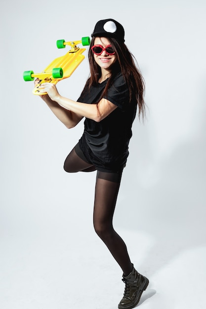 Feliz jovem morena em um top preto, shorts, meia-calça, boné e óculos de sol vermelhos com skate amarelo está posando em fundo branco no estúdio.