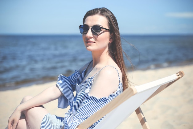 Feliz jovem morena relaxando em uma cadeira de deck de madeira na praia do oceano enquanto olha para a câmera, sorrindo e usando óculos escuros da moda. O conceito de férias desfrutando