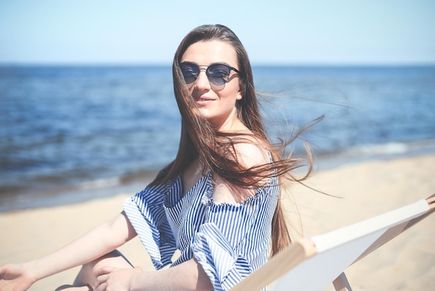 Feliz jovem morena relaxando em uma cadeira de deck de madeira na praia do oceano enquanto olha para a câmera, sorrindo e usando óculos escuros da moda. O conceito de férias desfrutando