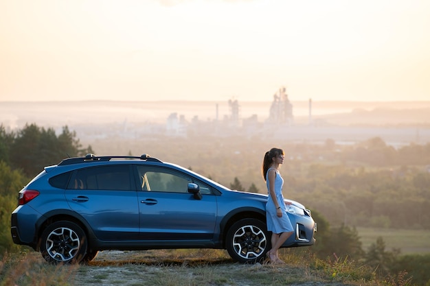 Feliz jovem motorista de vestido azul aproveitando a noite quente de verão ao lado de seu carro Conceito de viagem e férias