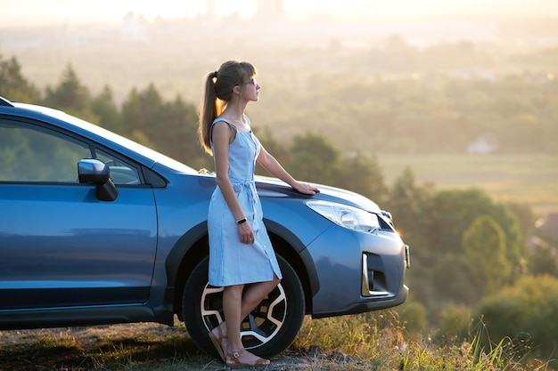 Feliz jovem motorista de vestido azul aproveitando a noite quente de verão ao lado de seu carro Conceito de viagem e férias