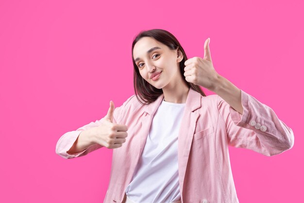 Feliz jovem mulher caucasiana em uma jaqueta rosa fazendo sinal de polegar para cima e sorrindo Bom trabalho e respeito