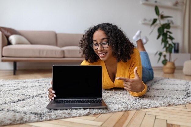 Feliz jovem negra deitada no chão em casa apontando para laptop com maquete para site em