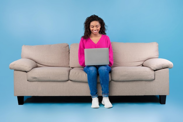 Feliz jovem negra em roupas casuais trabalhando online sentada no sofá e usando laptop em azul