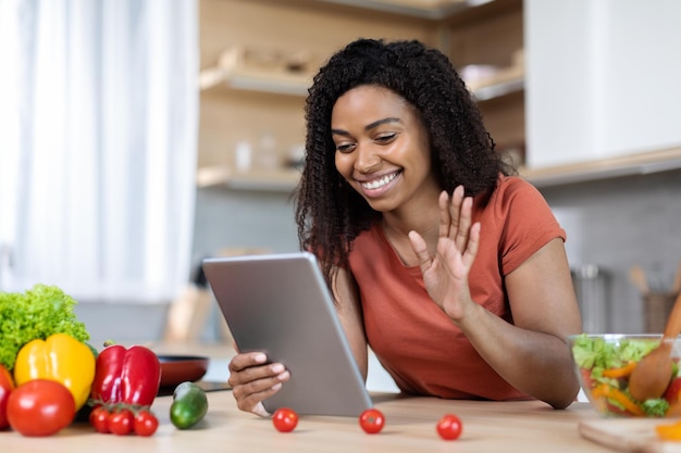 Feliz jovem negra em uma camiseta vermelha acenando para um tablet tem uma videochamada em uma mesa com legumes limitados