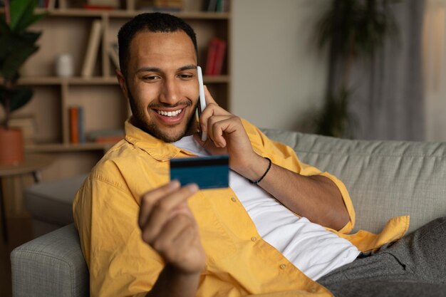 Feliz jovem negro com cartão de crédito fazendo pedidos de compras via smartphone em casa