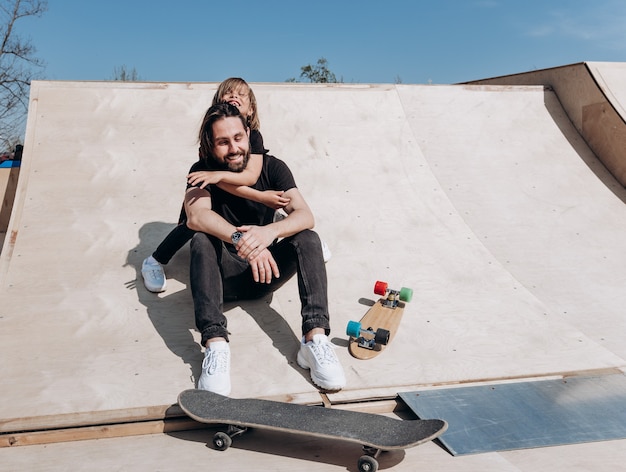 Feliz jovem pai e o filho vestido com roupas casuais elegantes estão sentados em um abraço juntos no slide ao lado dos skates em um parque de skate no dia quente e ensolarado.
