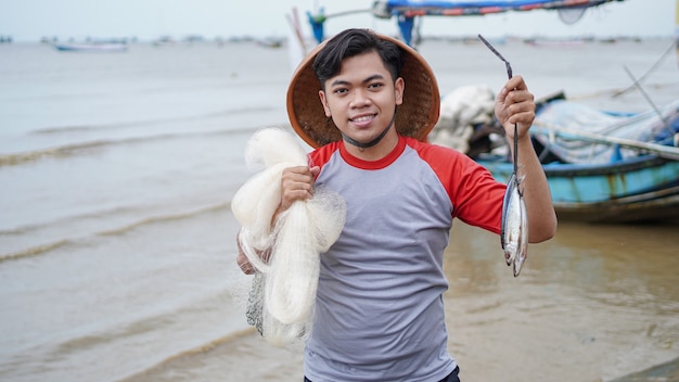 Feliz jovem pescador na praia segurando sua pescaria e shows na frente de seu barco