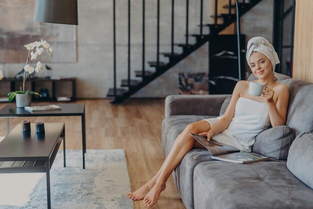 Foto feliz jovem posa envolta em toalha de banho bebe café aromático e lê revista senta-se no sofá confortável posa na sala de estar em casa atmoshpere doméstico estilo de vida de lazer