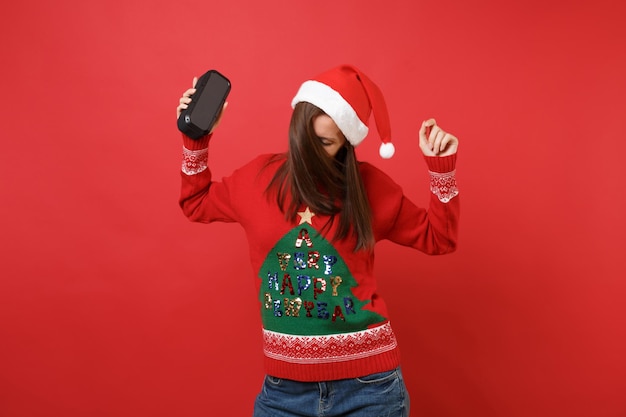 Feliz jovem santa dançando pulando, segurando o alto-falante portátil sem fio bluetooth música isolado em fundo vermelho brilhante. Conceito de festa de feriado de celebração de ano novo feliz 2019. Simule o espaço da cópia.