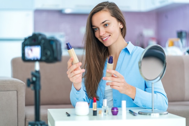 Feliz jovem sorridente mulher vídeo blogger influenciador segurando a fundação durante a gravação de beleza, maquiagem e cosméticos blog em casa.