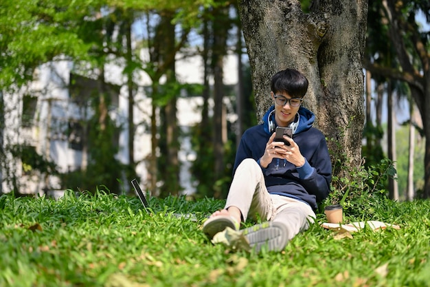 Feliz jovem universitário asiático usando seu smartphone debaixo da árvore no parque