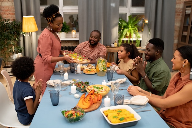 Feliz joven africana poniendo apetitosos sándwiches caseros para su familia en la mesa festiva entre velas encendidas y otros alimentos