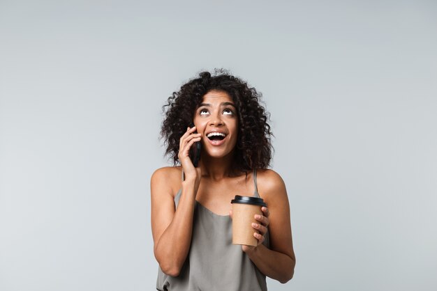 Feliz joven africana vestida casualmente que se encuentran aisladas, hablando por teléfono móvil, sosteniendo una taza de café para llevar