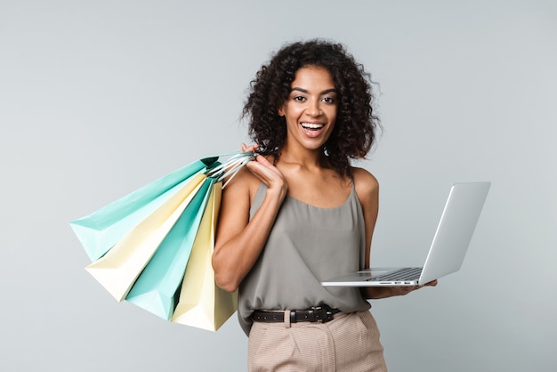 Feliz joven africana vestida casualmente que se encuentran aisladas, sosteniendo el ordenador portátil, llevando bolsas de la compra.