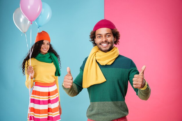 Feliz joven africano mostrando los pulgares para arriba mientras una niña sonriente con globos de pie