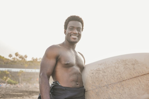 Feliz joven africano sonriendo a la cámara mientras sostiene una tabla de surf en la playa Concepto de deporte extremo