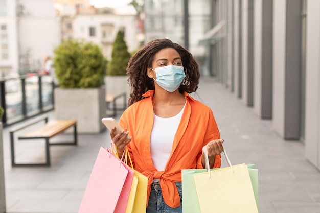 Feliz joven afroamericana con máscara protectora casual con muchas bolsas y teléfono inteligente