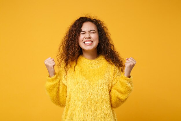 Feliz joven afroamericana con suéter de piel posando aislada en un fondo amarillo anaranjado, retrato de estudio. Concepto de estilo de vida de las personas. Simulacros de espacio de copia. Manteniendo los ojos cerrados, haciendo gesto de ganador.