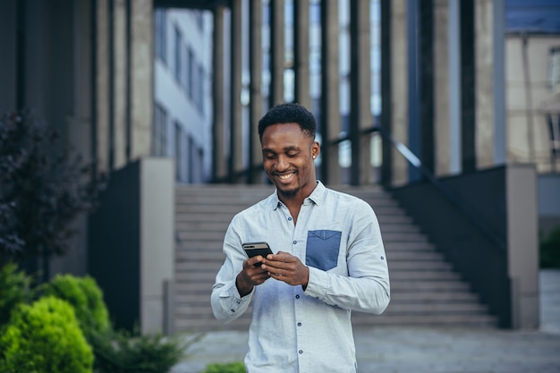 Feliz joven afroamericano independiente masculino correspondiente en el móvil y leyendo buenas noticias desde el teléfono móvil en línea