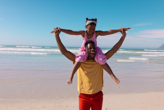 Feliz joven afroamericano tomados de la mano y llevando a la niña sobre los hombros contra el mar y el cielo