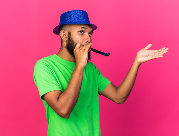 Feliz joven afroamericano vistiendo gorro de fiesta soplando silbato de fiesta extendiendo la mano