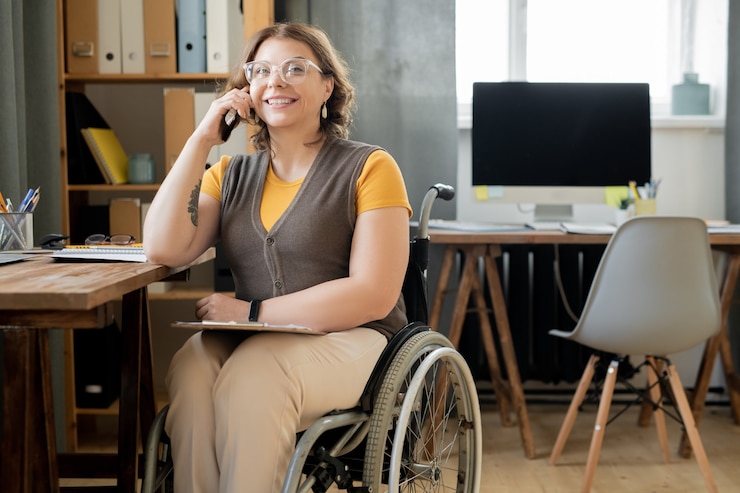 Foto feliz joven agente o secretaria en silla de ruedas sentada junto a la mesa en la oficina moderna y consultar a los clientes en el teléfono móvil por el lugar de trabajo