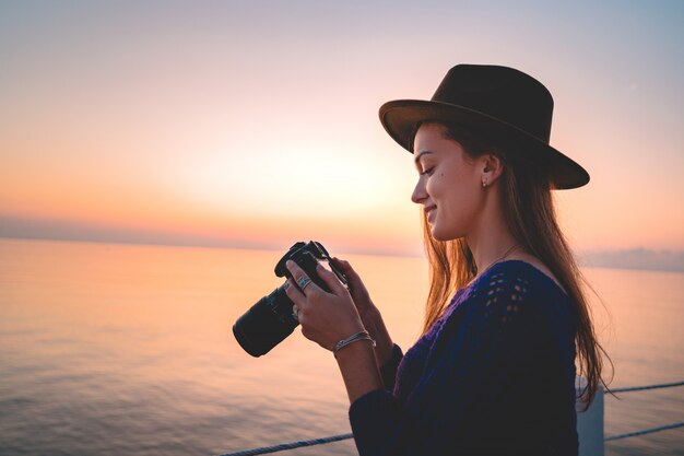 Feliz joven alegre fotógrafo viajero mujer con sombrero con cámara durante la toma de fotos del mar al atardecer