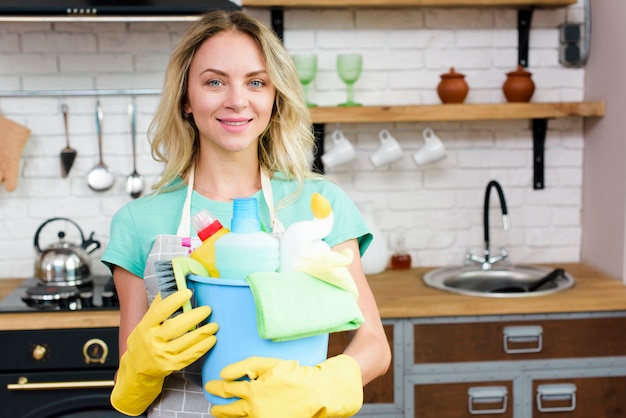 Feliz joven ama de casa de pie en la cocina con un cubo de productos de limpieza