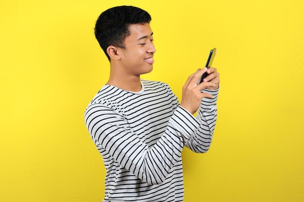 Feliz joven apuesto hombre asiático sonriendo con smartphone aislado sobre fondo amarillo