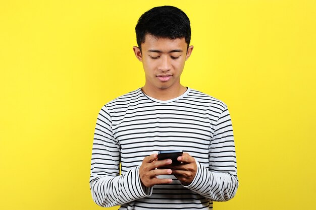 Feliz joven apuesto hombre asiático sonriendo con smartphone aislado sobre fondo amarillo