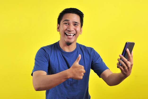 Feliz de joven apuesto hombre asiático sonriendo con smartphone aislado sobre fondo amarillo