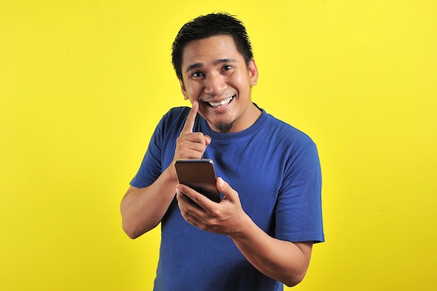 Feliz joven apuesto hombre asiático sonriendo con teléfono móvil, aislado sobre fondo amarillo