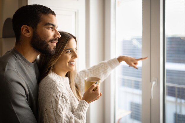 Feliz joven árabe abraza a la esposa europea dama señalando con el dedo por la ventana y bebe una taza de té