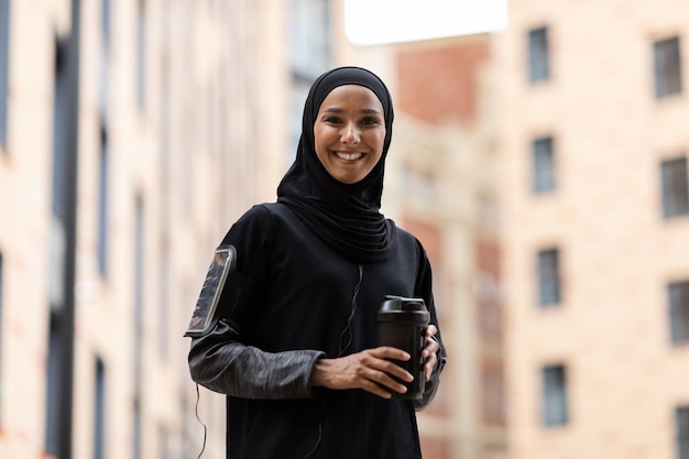 Feliz joven árabe con hiyab y uniforme deportivo con teléfono en el hombro con una taza de cóctel