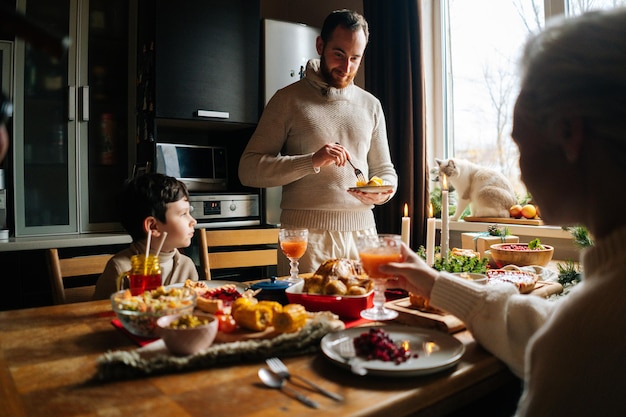 Feliz joven arreglando comida en platos para esposa e hijo de pie en la mesa festiva de Navidad durante