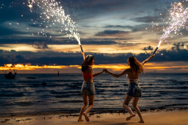 Feliz joven asiática bailando y jugando a los fuegos artificiales con chispas juntos en la playa de una isla tropical en la noche de verano chica atractiva disfrutar y divertirse al aire libre estilo de vida viaje océano en vacaciones vacaciones