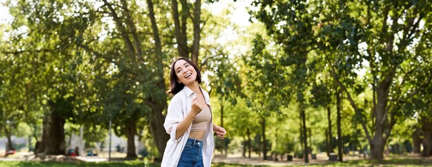 Feliz joven asiática caminando sola bailando y cantando en el parque sonriendo gente despreocupada y