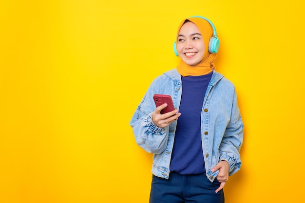 Feliz joven asiática con chaqueta de jeans y auriculares sosteniendo un teléfono móvil mirando a un lado el espacio de copia aislado sobre el fondo amarillo