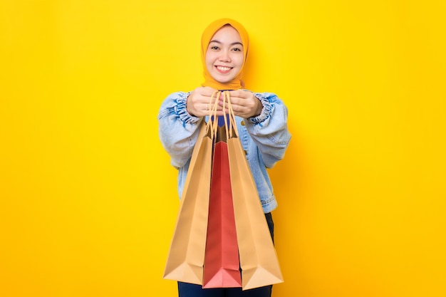 Feliz joven asiática con chaqueta de jeans sosteniendo bolsas de compras y sonriendo a la cámara aislada sobre el fondo amarillo