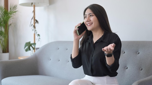 Foto feliz joven asiática descansando en el sofá en casa y hablando por teléfono celular