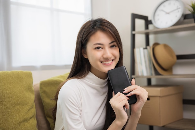 Feliz joven asiática mirando la pantalla del teléfono inteligente sentada en el sofá en la sala de estar comprando en línea Ella muy feliz Adolescente leyendo noticias del teléfono móvil sintiéndose emocionada