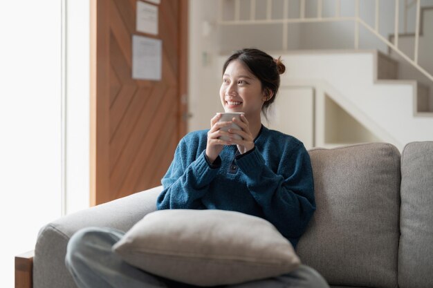 Feliz joven asiática sentada en un sofá con una taza de café en casa