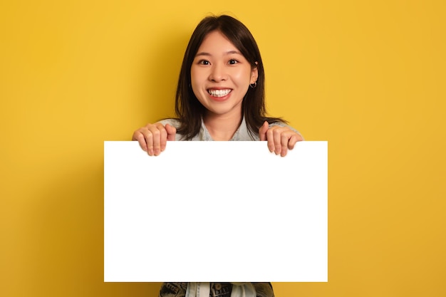 Feliz joven asiática sosteniendo una pancarta de papel en blanco sonriendo a la cámara en el fondo amarillo del estudio