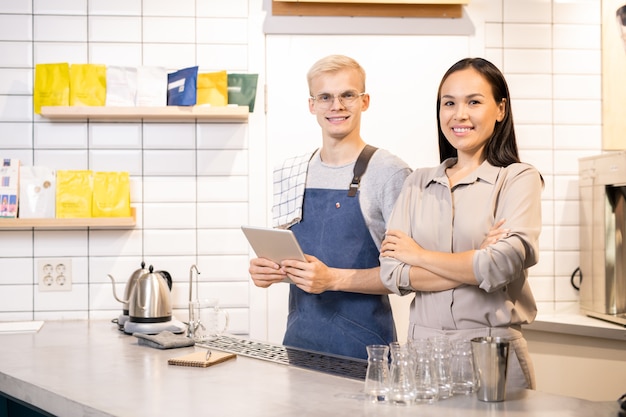 Feliz joven asiática y su colega masculino en ropa de trabajo mirándote con sonrisas amistosas durante el trabajo