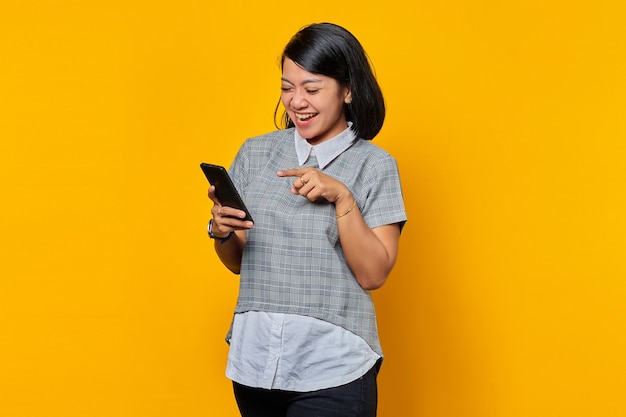 Feliz joven asiática usando un teléfono móvil y mirando la pantalla del teléfono inteligente sobre fondo amarillo