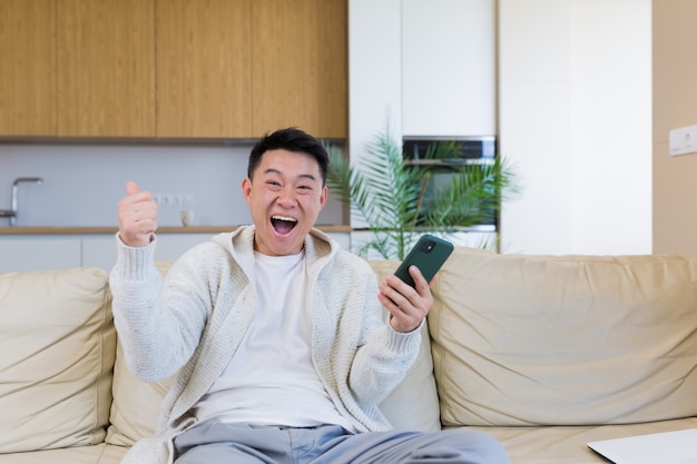 Feliz joven asiático en casa mirando celular con emoción de ganador