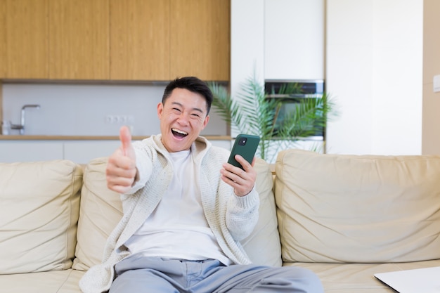 Feliz joven asiático en casa mirando celular con emoción de ganador