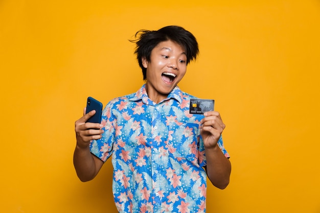 Feliz joven asiático que se encuentran aisladas sobre el espacio amarillo mediante teléfono móvil con tarjeta de crédito.